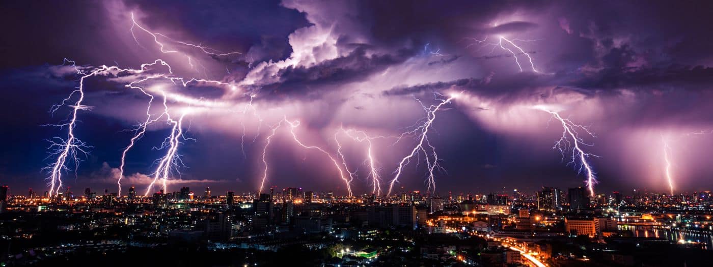 Di notte, numerosi fulmini colpiscono il profilo di una città, illuminando nuvole spettacolari e proiettando un bagliore viola sul paesaggio urbano sottostante.