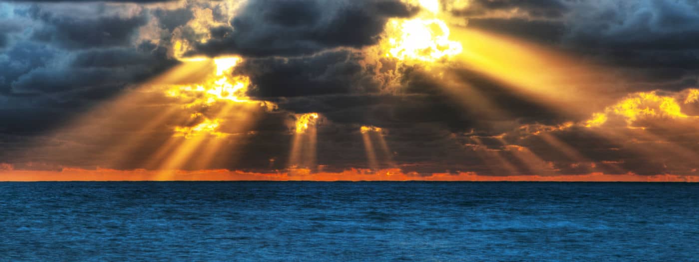 I raggi del sole filtrano attraverso le nuvole scure sopra un oceano blu all'alba o al tramonto, creando un bagliore dorato all'orizzonte.