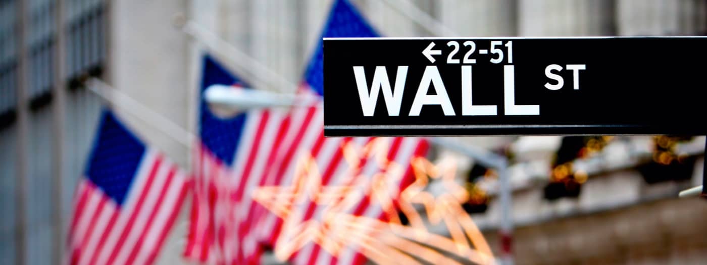 Un cartello di Wall Street con i numeri 22-51 è esposto in modo evidente. Sullo sfondo, diverse bandiere americane sono leggermente sfocate, enfatizzando l'atmosfera del distretto finanziario.