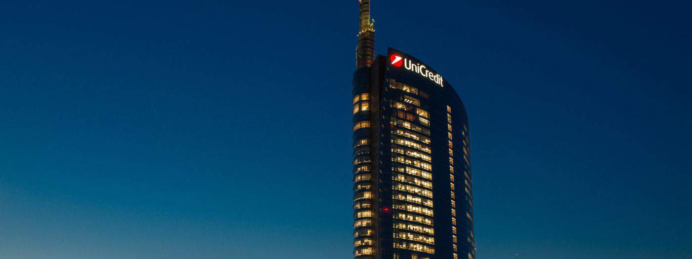 Un grattacielo alto e moderno con architettura curva e finestre illuminate si staglia contro un cielo notturno limpido. L'edificio presenta un logo in cima con il testo "UniCredit.