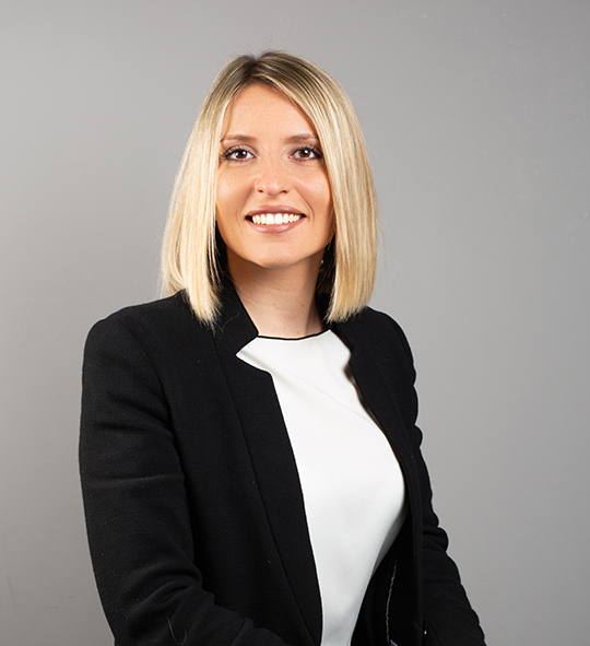 Una persona con i capelli biondi lunghi fino alle spalle sorride mentre indossa un blazer nero sopra una maglietta bianca su uno sfondo grigio.