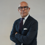 Un uomo calvo con occhiali, abito scuro, camicia bianca e cravatta viola sta in piedi con le braccia incrociate su uno sfondo chiaro, guardando direttamente la telecamera.