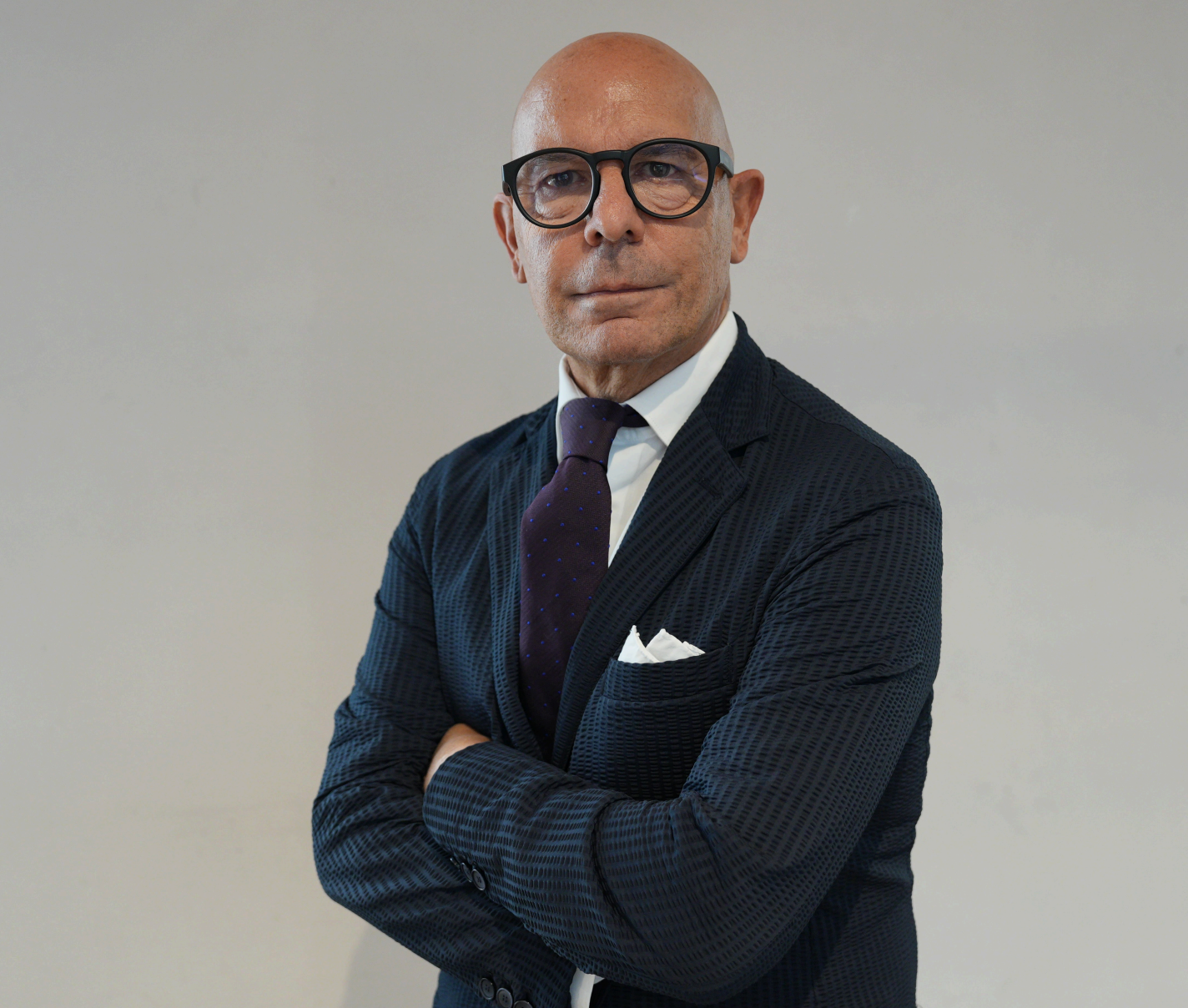 Un uomo calvo con occhiali, abito scuro, camicia bianca e cravatta viola sta in piedi con le braccia incrociate su uno sfondo chiaro, guardando direttamente la telecamera.