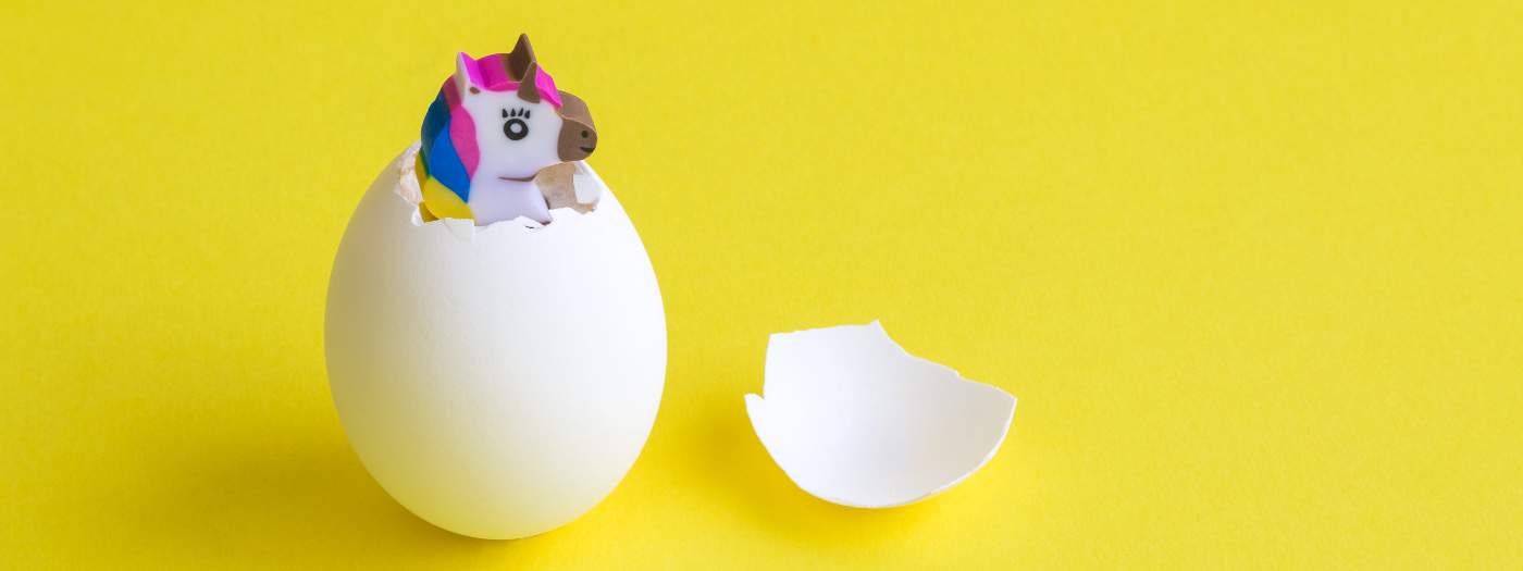 Un unicorno giocattolo con i capelli colorati emerge da un uovo bianco rotto su uno sfondo giallo brillante. I pezzi di guscio dell'uovo sono adiacenti all'uovo intero.