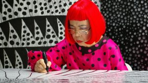 Donna con parrucca rossa e abito a pois rosa disegna con un pennarello nero su un tavolo.