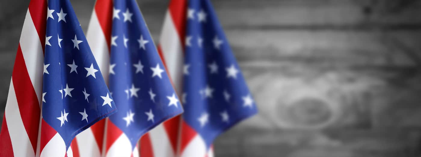 Quattro bandiere americane a stelle e strisce sono allineate in primo piano, con uno sfondo di legno grigio sfocato alle loro spalle.
