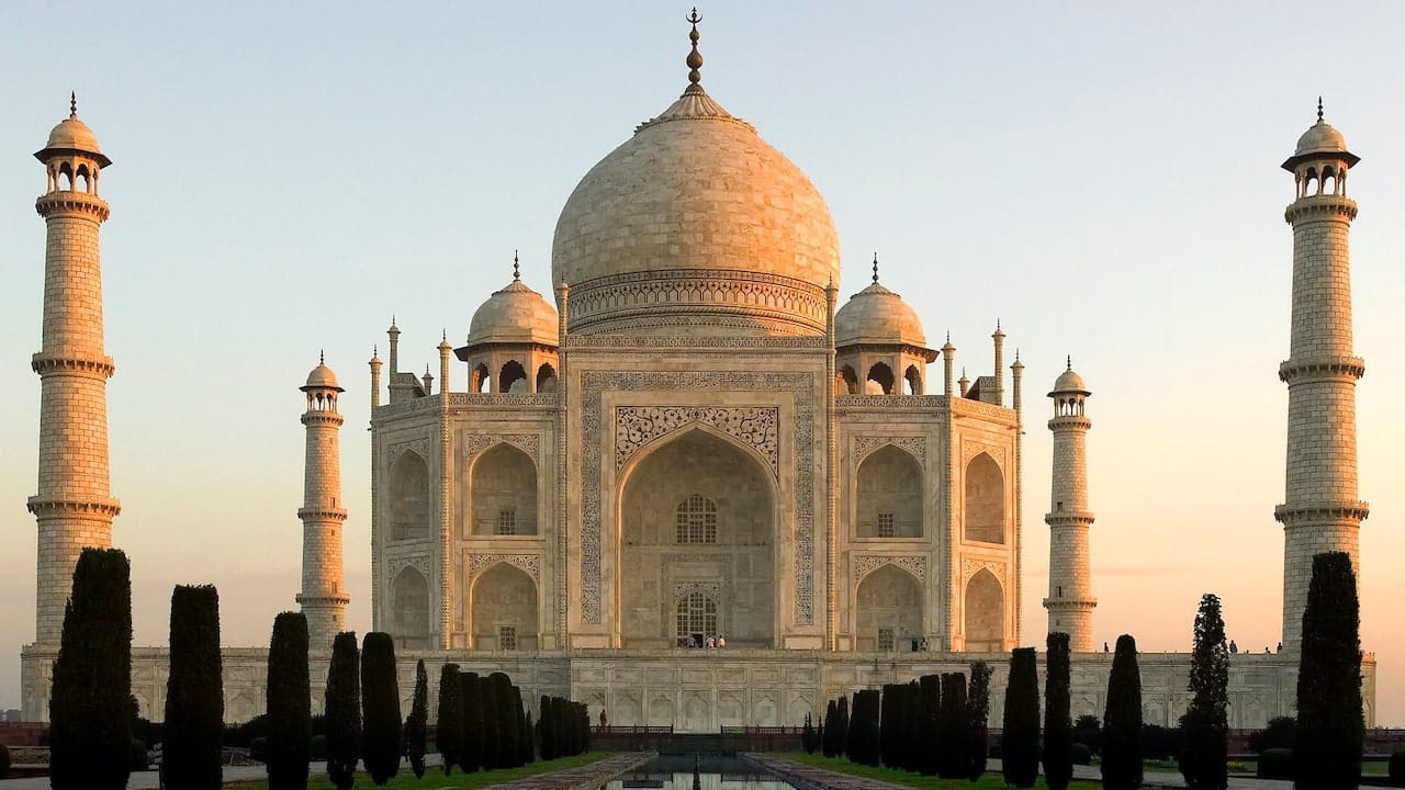 Il Taj Mahal, un mausoleo di marmo bianco con una grande cupola centrale, quattro minareti e giardini simmetrici, si erge sotto un cielo limpido al tramonto.