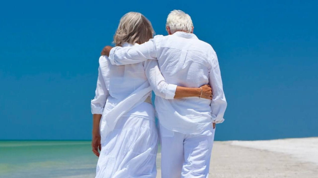 Una coppia di anziani vestiti di bianco cammina a braccetto lungo una spiaggia sabbiosa, con l'oceano e il cielo azzurro e limpido sullo sfondo.