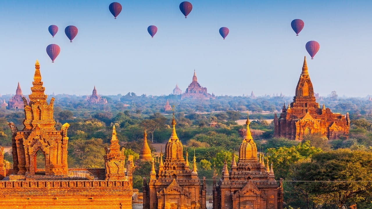 Mongolfiere che sorvolano un antico paesaggio di templi con vegetazione lussureggiante e guglie architettoniche durante una giornata limpida a Bagan, Myanmar. Il cielo è azzurro, offrendo uno sfondo scenografico al sito storico.
