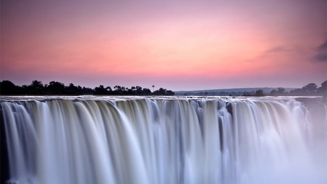Una maestosa cascata precipita da un'ampia rupe, con acqua liscia come la seta catturata in movimento. Il cielo sopra è dipinto con tenui tonalità di rosa e viola, che indicano l'alba o il tramonto. Una vegetazione lussureggiante delimita l'orizzonte sullo sfondo.