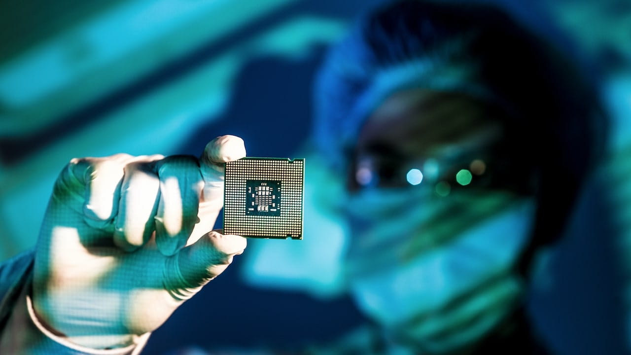 Una persona con indumenti protettivi e guanti tiene in mano un microchip, con uno sfondo sfocato che suggerisce un ambiente high-tech o di laboratorio.