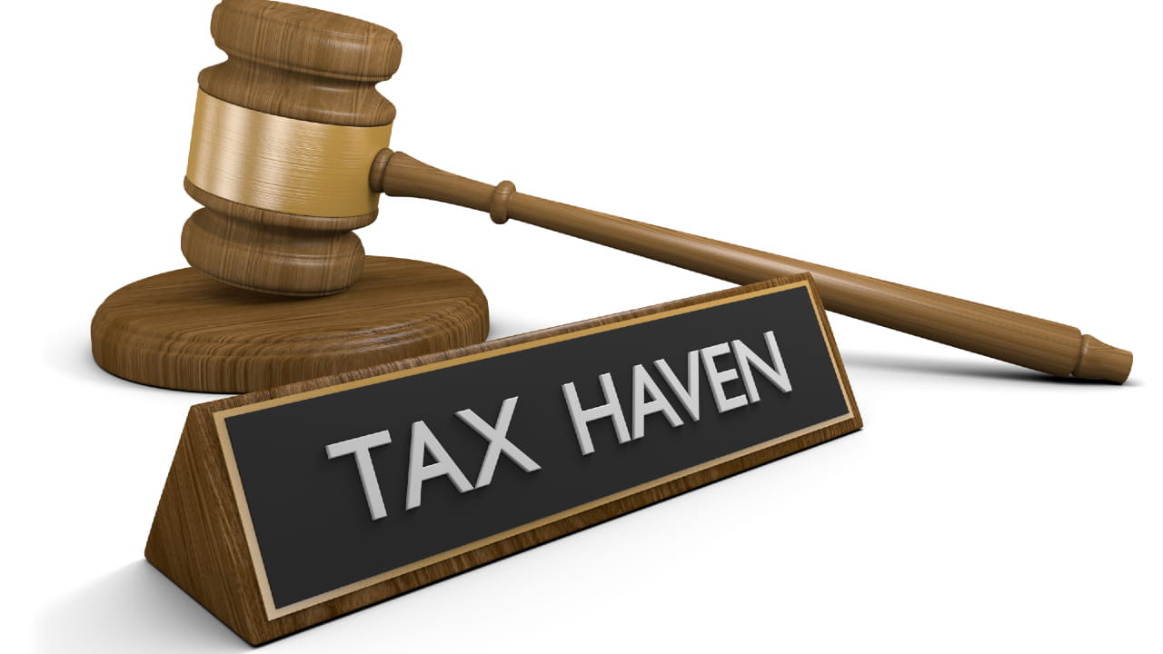 Un martelletto da giudice in legno è appoggiato accanto a una targhetta con la scritta "Tax haven" (paradiso fiscale) in caratteri bianchi su sfondo nero, a sottolineare il controllo che tali giurisdizioni devono affrontare da parte di organizzazioni come l'Ocse.