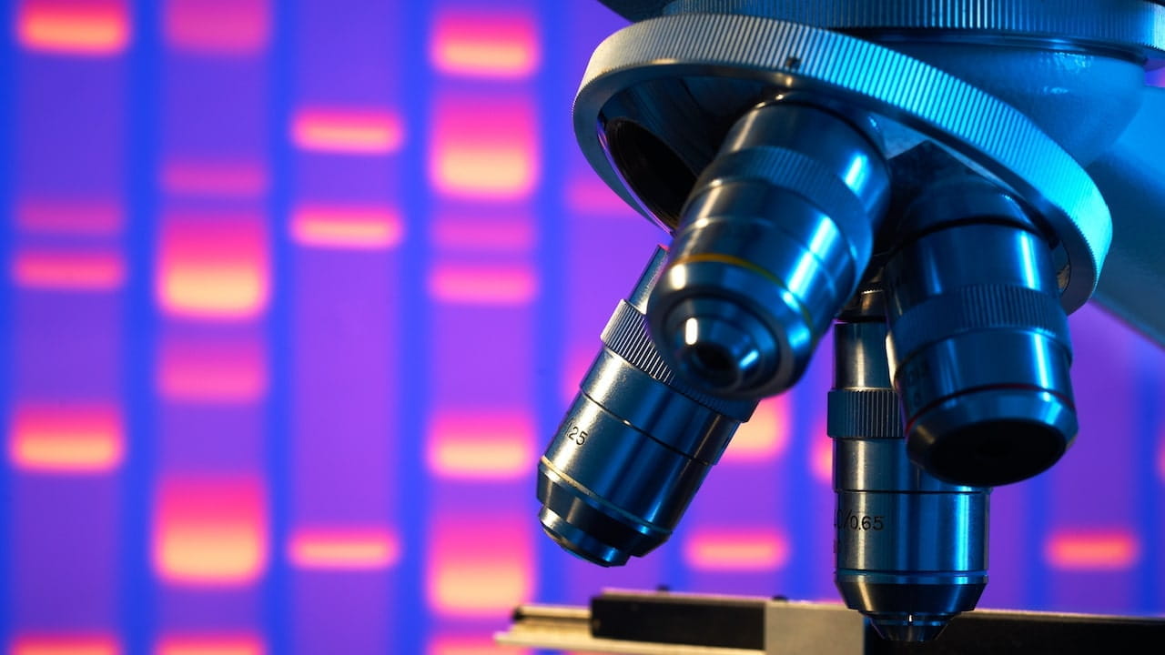 Primo piano delle lenti dell'obiettivo di un microscopio con sfondo sfocato che mostra bande di DNA colorate, a suggerire un contesto di ricerca scientifica o genetica.