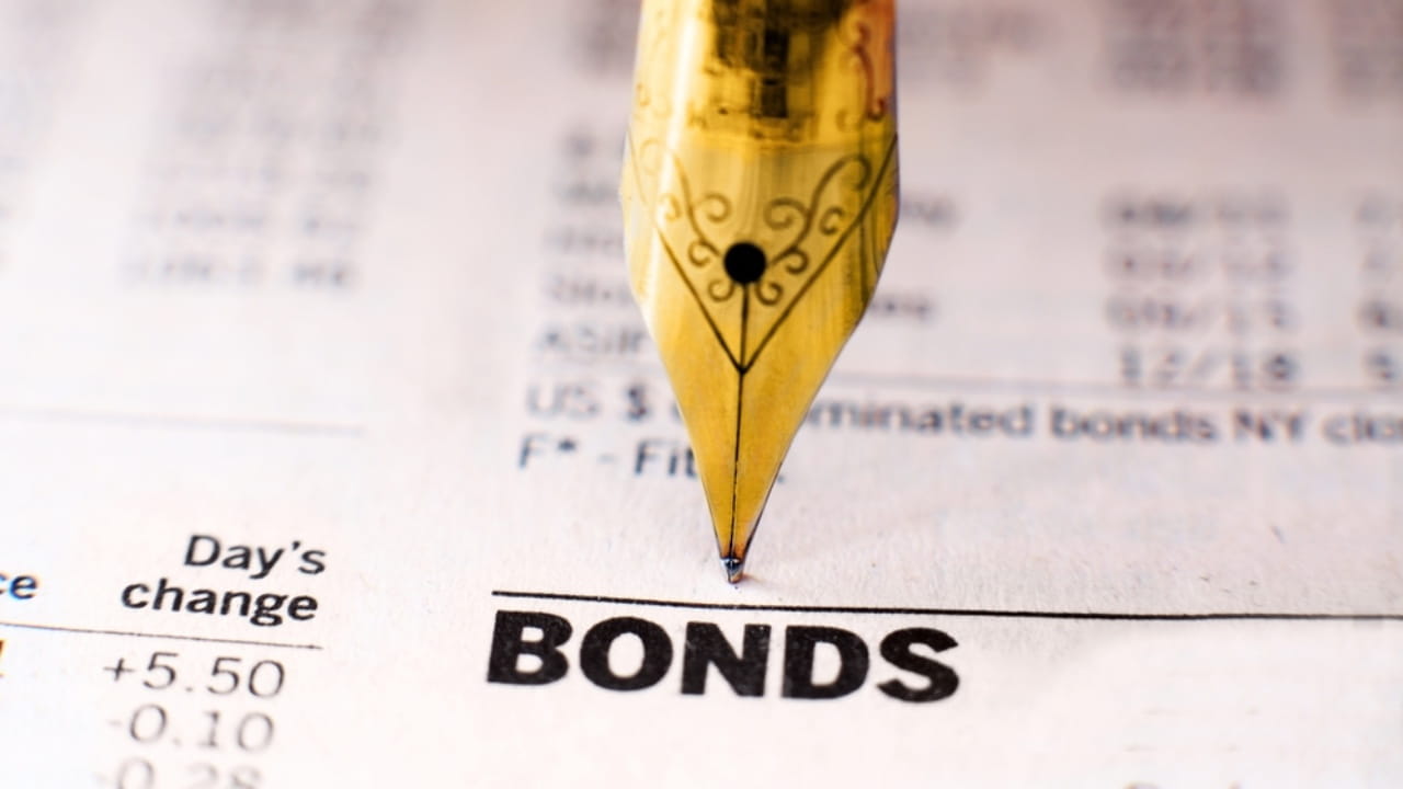 Primo piano del pennino di una penna stilografica appoggiato su una pagina di giornale finanziario con la parola "BONDS" in bella vista. La pagina include vari dati e cifre finanziarie.