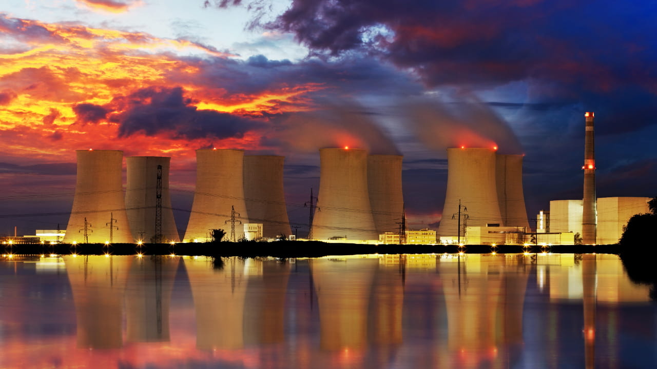 Una centrale nucleare con torri di raffreddamento multiple emette vapore al tramonto, riflettendosi nell'acqua sottostante. Il cielo è drammatico con nuvole arancioni, viola e blu. I tralicci dell'elettricità sono visibili in primo piano.