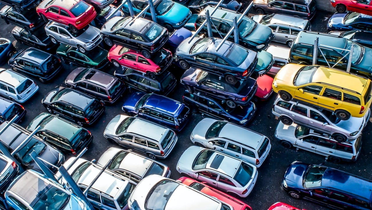 Una vista aerea di un affollato deposito di rottami con pile di auto di vari colori, tra cui rosso, blu, bianco e giallo. Le auto sono stipate fittamente, creando un aspetto organizzato ma caotico.