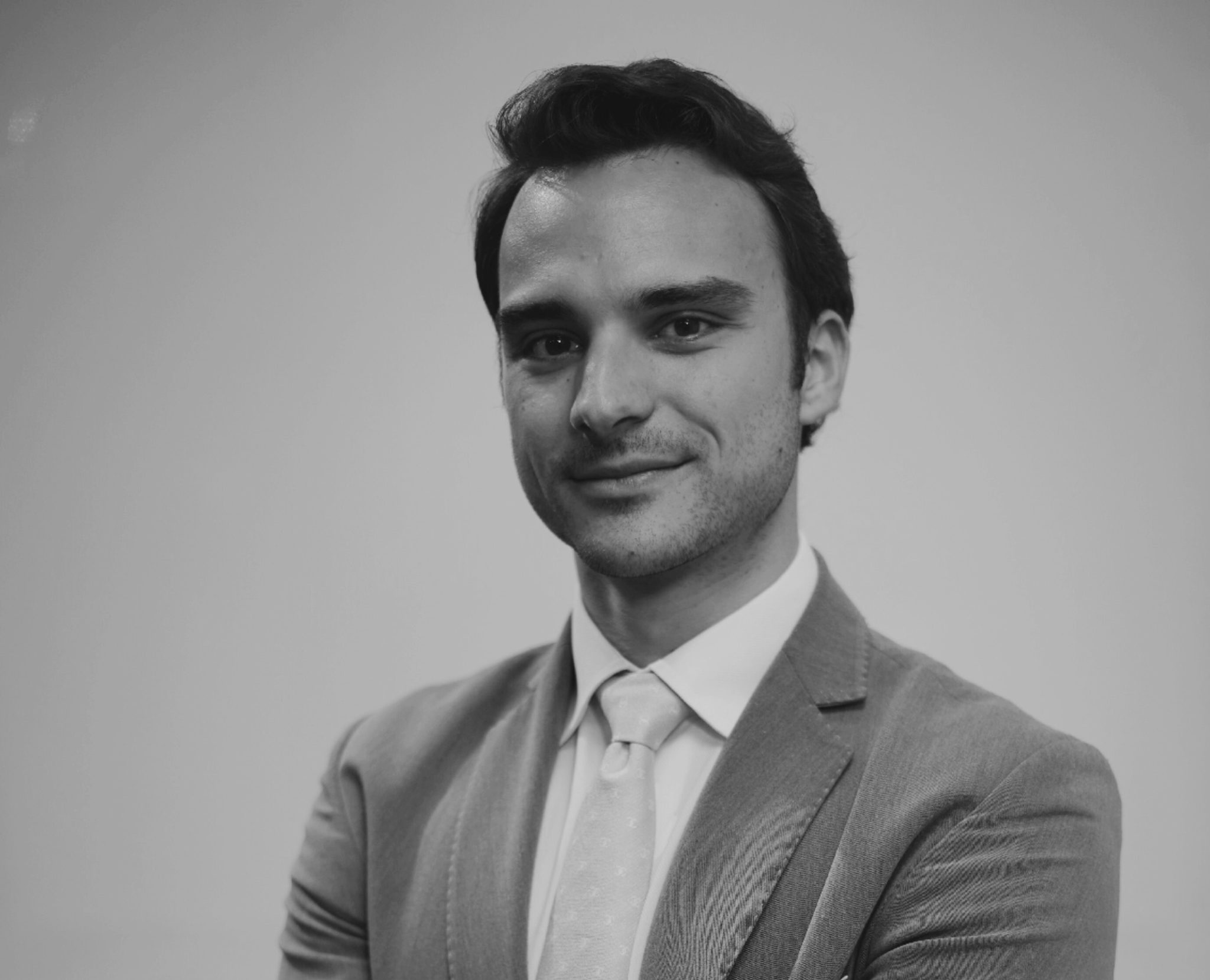 A man in a suit and tie stands with arms crossed, looking confidently at the camera. The image is in black and white with a plain background.