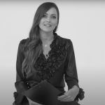 A woman with long, wavy hair wearing a ruffled blouse sits and smiles while holding a clipboard against a plain, light background. The image is in black and white.