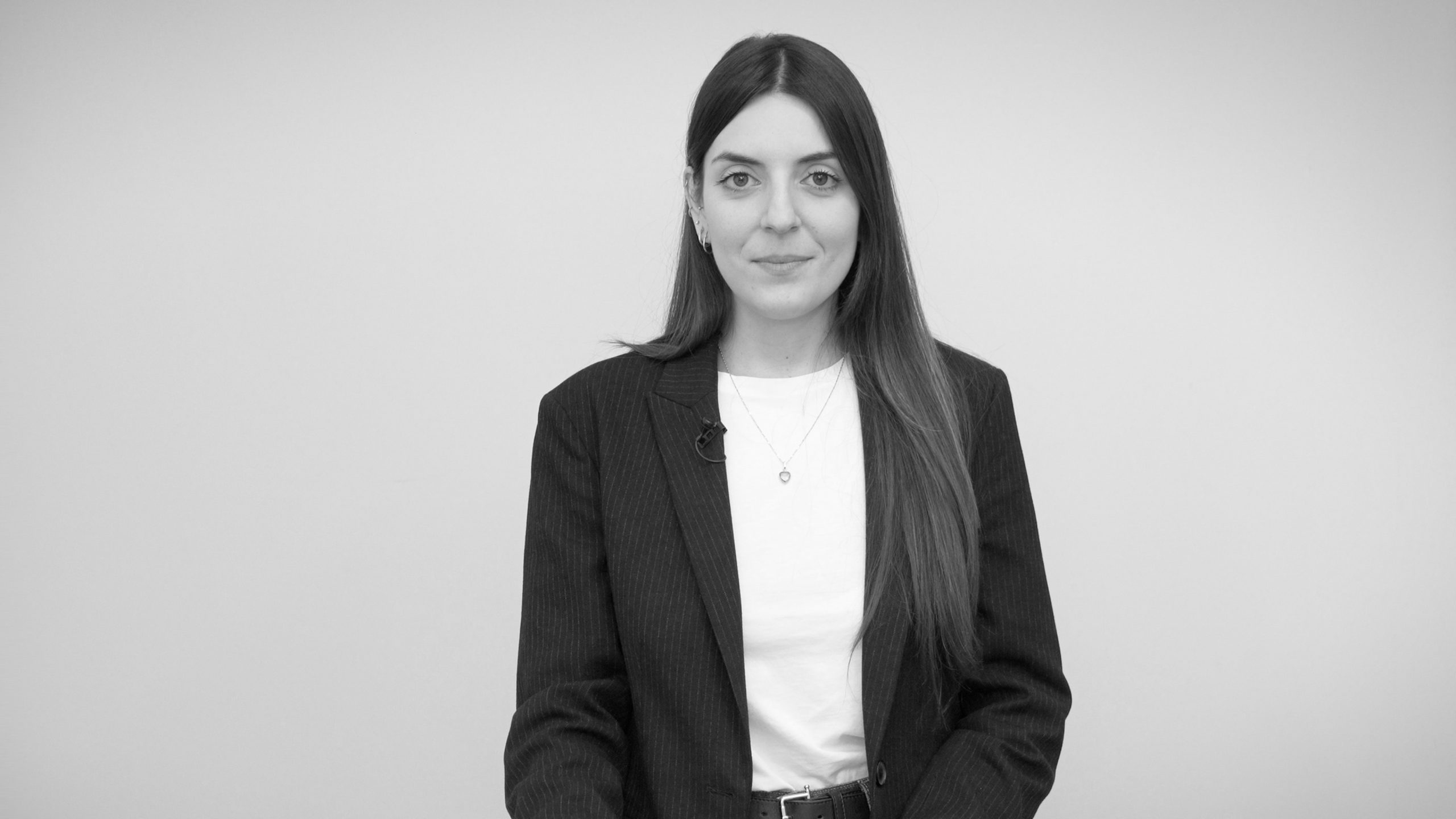 A young woman with long brown hair, wearing a dark blazer and white shirt, stands against a plain light background, looking at the camera and smiling slightly.