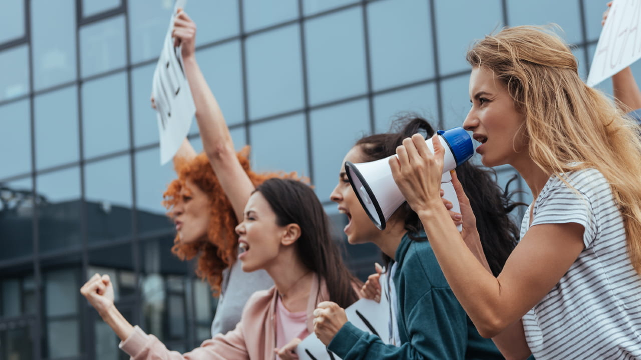 Un gruppo di persone protesta con passione di fronte a un moderno edificio in vetro. Tengono cartelli e una persona usa un megafono. Il gruppo sembra determinato, con le mani alzate e un'espressione di urgenza.