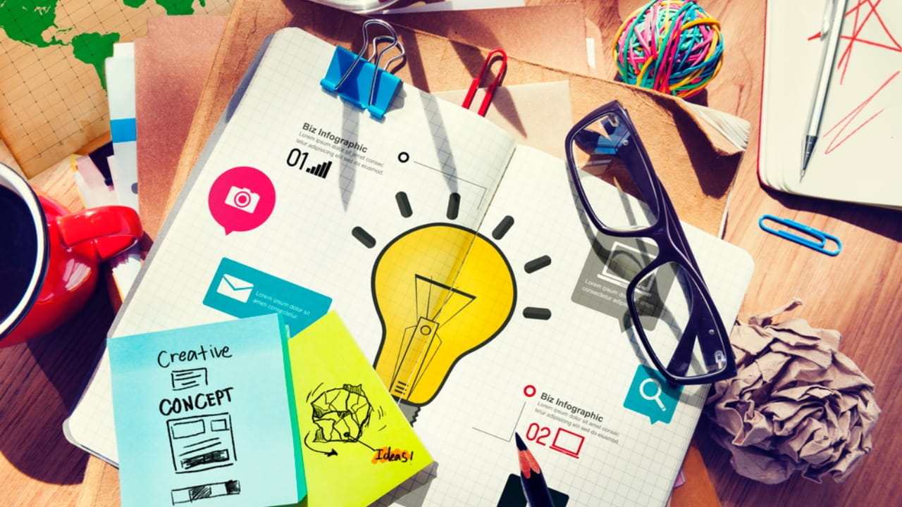 Una scrivania ingombra coperta da un quaderno con scarabocchi, grafici e un'illustrazione di una lampadina. Gli oggetti circostanti includono occhiali, graffette colorate, post-it, una palla di carta spiegazzata, una tazza rossa e un angolo del globo.