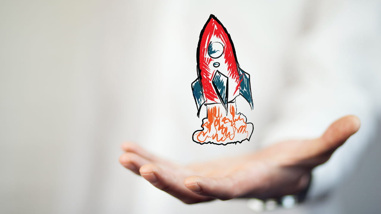 Una mano è tenuta con il palmo rivolto verso l'alto con un'illustrazione di un razzo rosso che decolla, lasciando dietro di sé fiamme arancioni e fumo. Lo sfondo è sfocato, enfatizzando il razzo e la mano aperta.