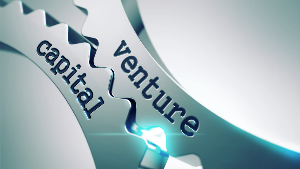 Close-up image of two interlocking metallic gears, one labeled "venture" and the other labeled "capital," symbolizing the concept of venture capital and its interconnected components.