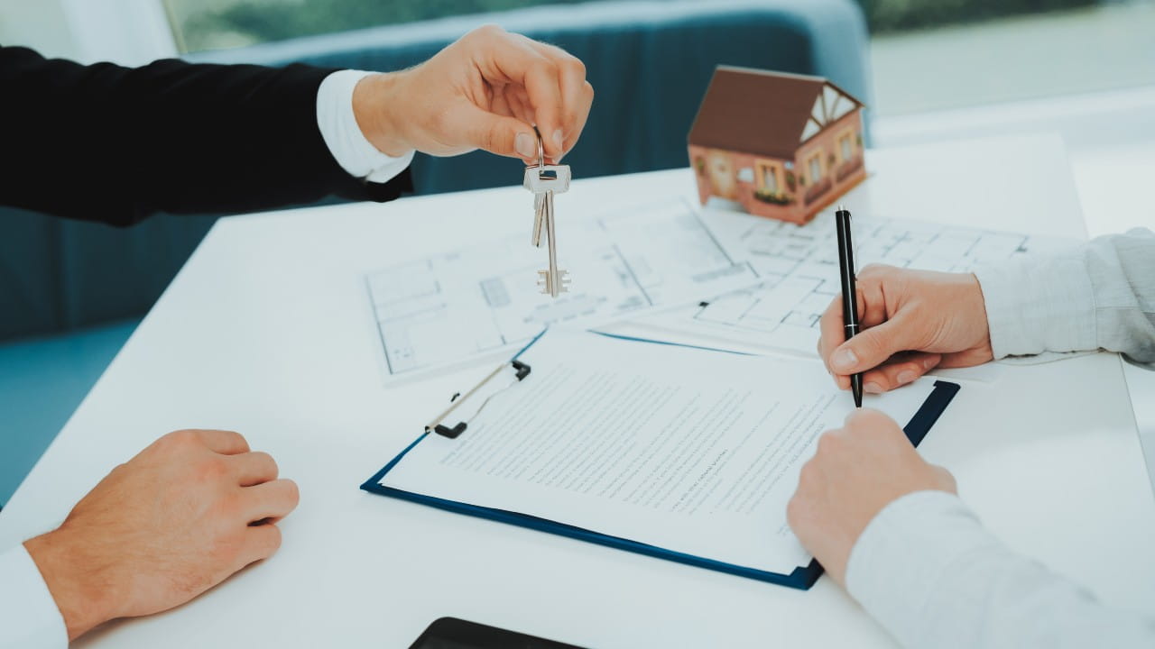 Due persone sono sedute a un tavolo con documenti immobiliari. Una consegna le chiavi all'altra, che sta firmando dei documenti. Sullo sfondo si vedono un piccolo modello di casa e planimetrie di un edificio, a suggerire una transazione immobiliare o un cambio di residenza.