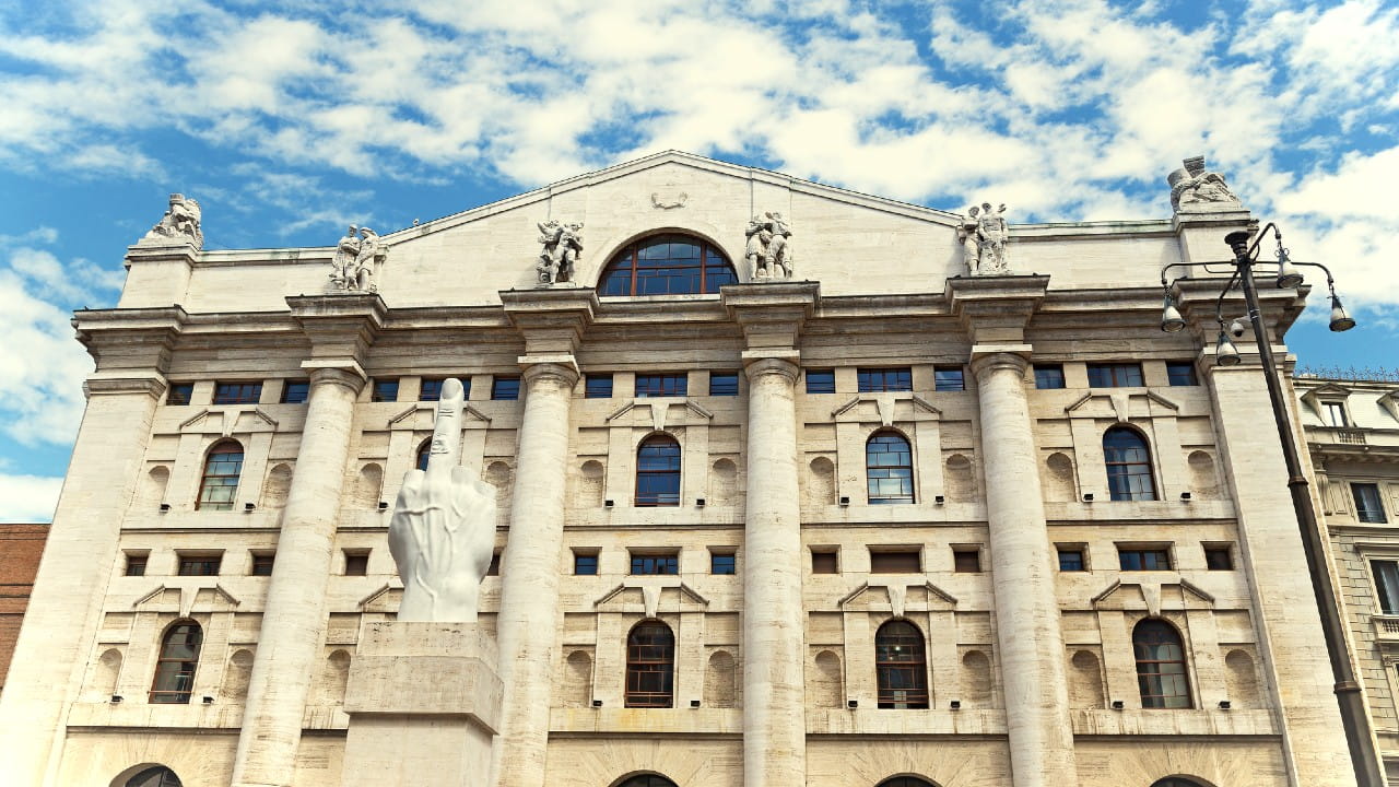 Un grande edificio neoclassico con alte colonne e statue sul tetto, con una grande scultura di mano con il dito medio allungato davanti, sotto un cielo azzurro parzialmente nuvoloso.