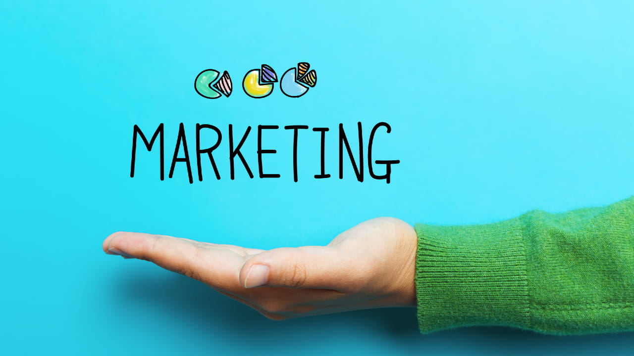 Una mano con una manica verde sostiene la parola "Marketing". Sopra la parola, ci sono forme astratte colorate su uno sfondo blu brillante.