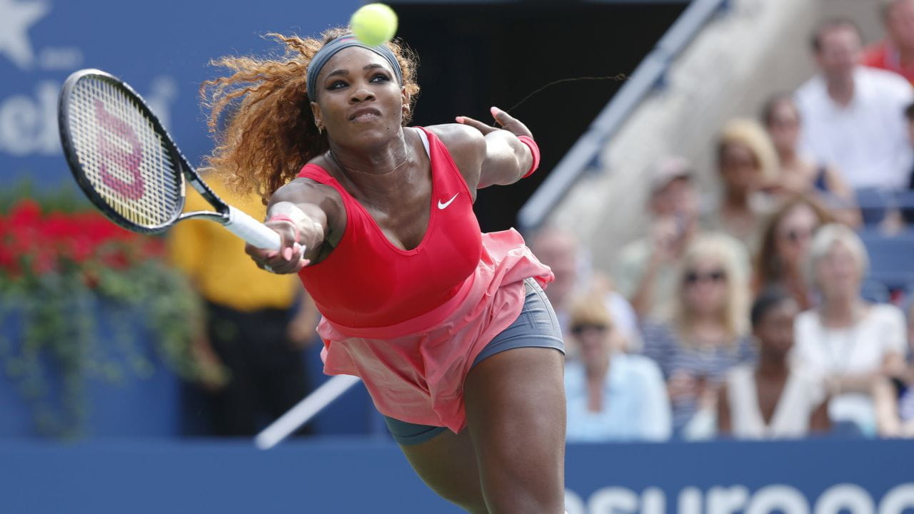 Una tennista vestita di rosso si lancia in avanti con la racchetta per colpire una pallina da tennis durante una partita, sfruttando la forza di Serena Williams, mentre una folla di spettatori osserva sullo sfondo.