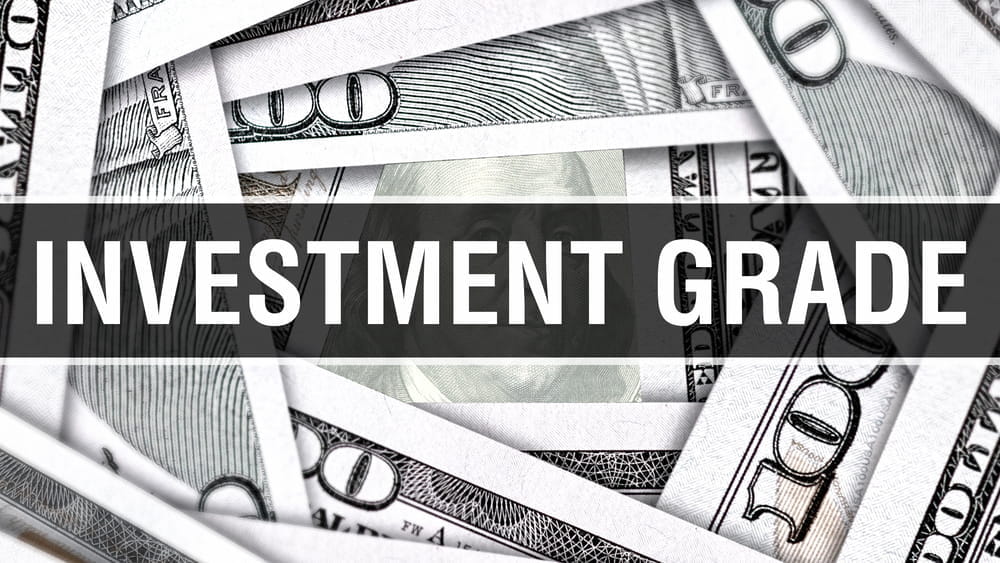 L'immagine mostra diverse banconote da cento dollari statunitensi sovrapposte con la scritta "INVESTMENT GRADE" scritta in grassetto e bianca al centro su una striscia scura trasparente.