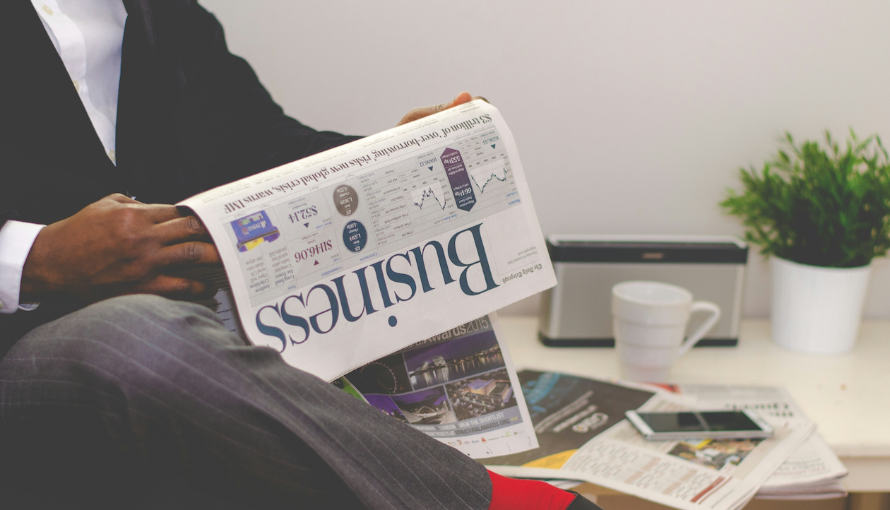 Una persona in giacca e cravatta siede e legge un giornale "Business", con particolare attenzione alla crescita consulenziale. Su un tavolo bianco vicino ci sono una tazza, un piattino, uno smartphone, documenti, una pianta e una radio, in un ambiente da ufficio o da salotto.