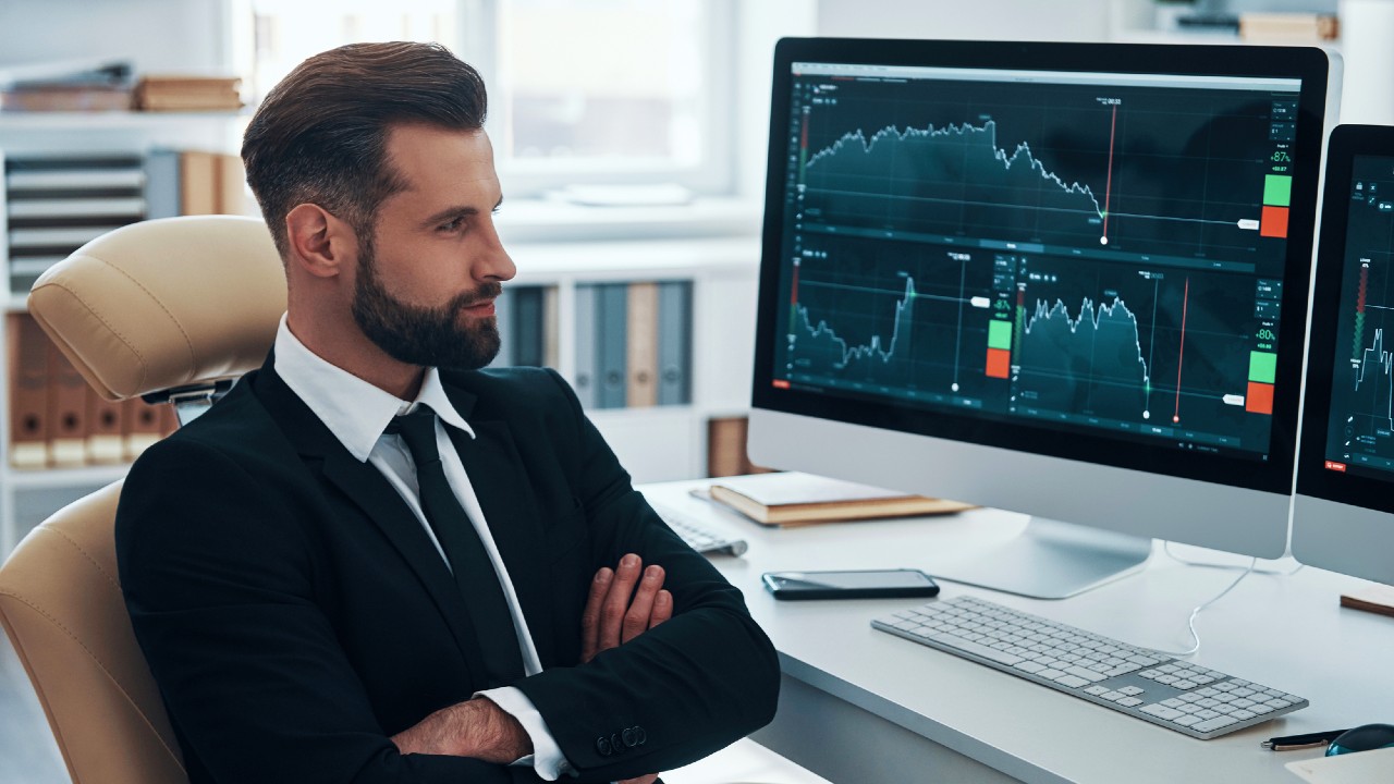 Un uomo in giacca e cravatta è seduto a una scrivania con le braccia incrociate, esaminando attentamente i monitor dei computer che mostrano grafici finanziari e grafici S&P 500. L'ambiente dell'ufficio comprende scaffali e una finestra sullo sfondo, che riflettono un'atmosfera concentrata.