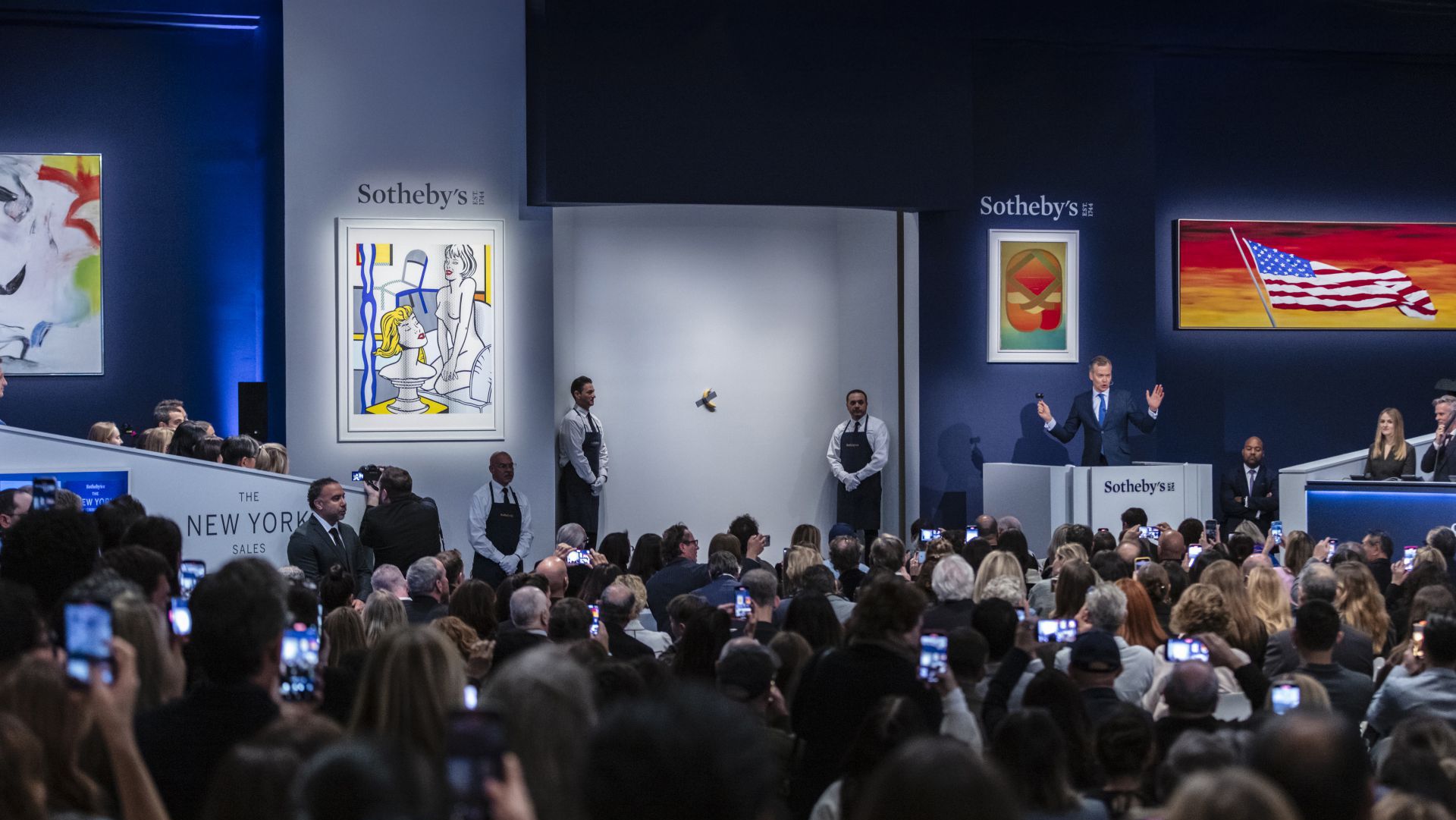 La sala d'aste affollata di Sotheby's brulica di partecipanti che catturano momenti sui loro telefoni. Un banditore tiene banco sul podio vicino a un grande schermo che mostra opere d'arte costose 2024, mentre due dipinti colorati adornano le pareti. Il personale in gilet scuri è lì vicino, attento.