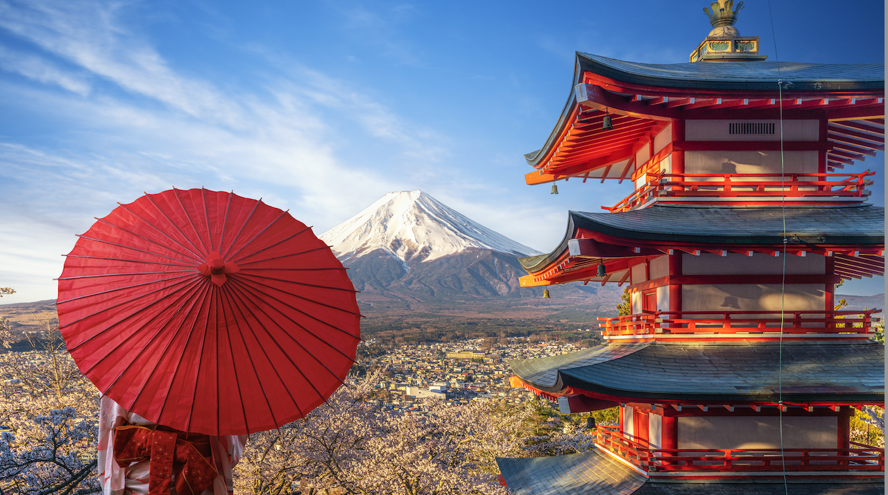 Una persona tiene un parasole rosso di fronte al Monte Fuji, incarnando l'essenza serena del Giappone. In primo piano, una pagoda rossa e bianca a cinque piani è circondata da ciliegi in fiore sotto un cielo blu vibrante.