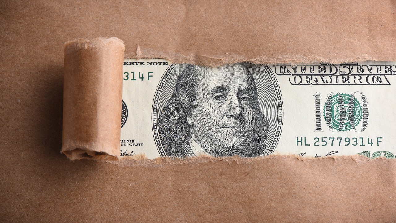Una banconota statunitense da cento dollari viene parzialmente svelata sotto una carta marrone strappata, mostrando il volto di Benjamin Franklin e parte del disegno di sicurezza OCSE della banconota.