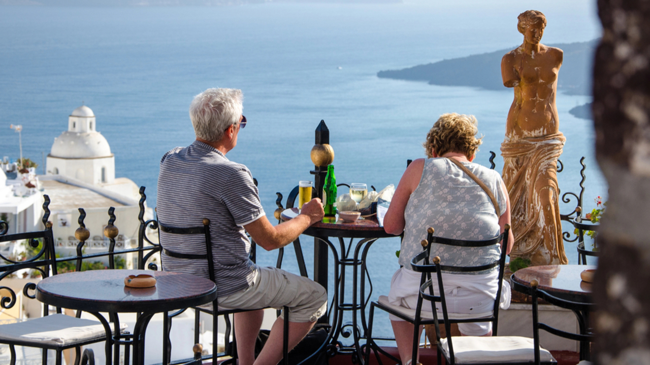 Una coppia è seduta a un tavolo su un balcone che si affaccia sul mare, discutendo dell'ultima riforma pensioni mentre si gode un drink e un piatto di cibo. Una statua si trova lì vicino, e edifici bianchi più una chiesa con una cupola abbelliscono lo sfondo contro l'acqua blu scintillante.