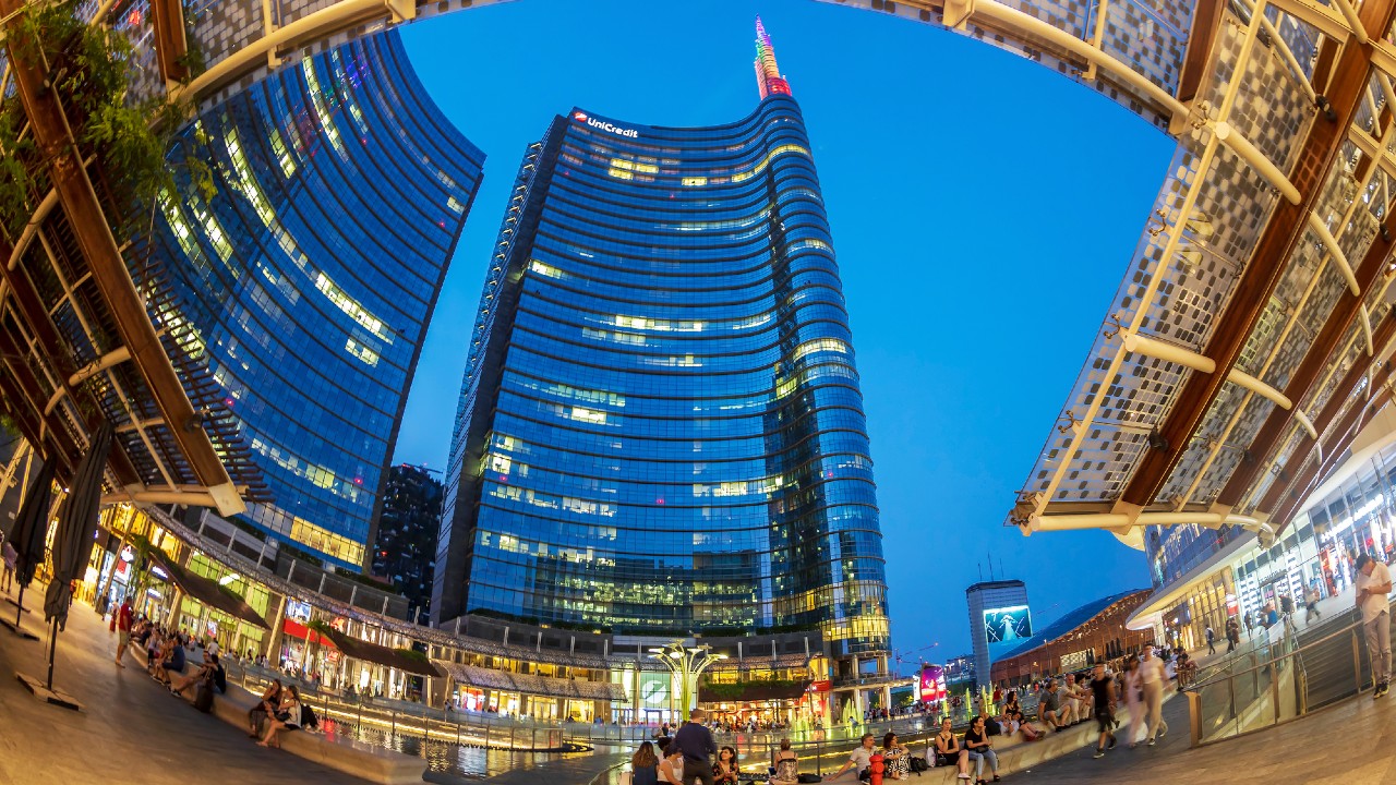 Vista fisheye di una vivace piazza urbana circondata da moderni grattacieli, tra cui l'iconica UniCredit Tower, sotto un limpido cielo serale. Le persone si radunano attorno a una fontana con l'edificio più alto al centro, illuminato da luci calde. Una tettoia copre parzialmente la scena.