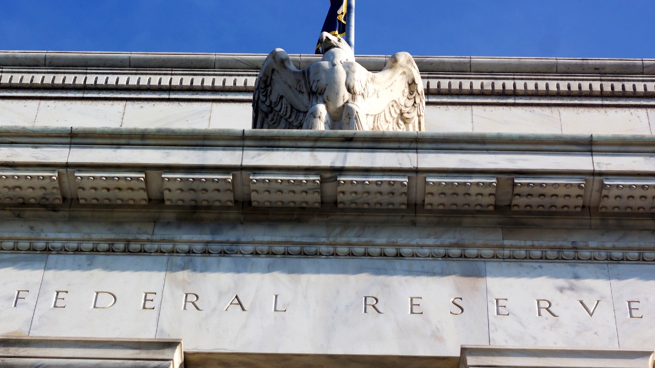 Vista dal basso della facciata di un edificio in pietra con la scritta "Federal Reserve" incisa sul muro. Una scultura di un'aquila è appollaiata su una sporgenza, con una piccola porzione di cielo azzurro visibile sullo sfondo.