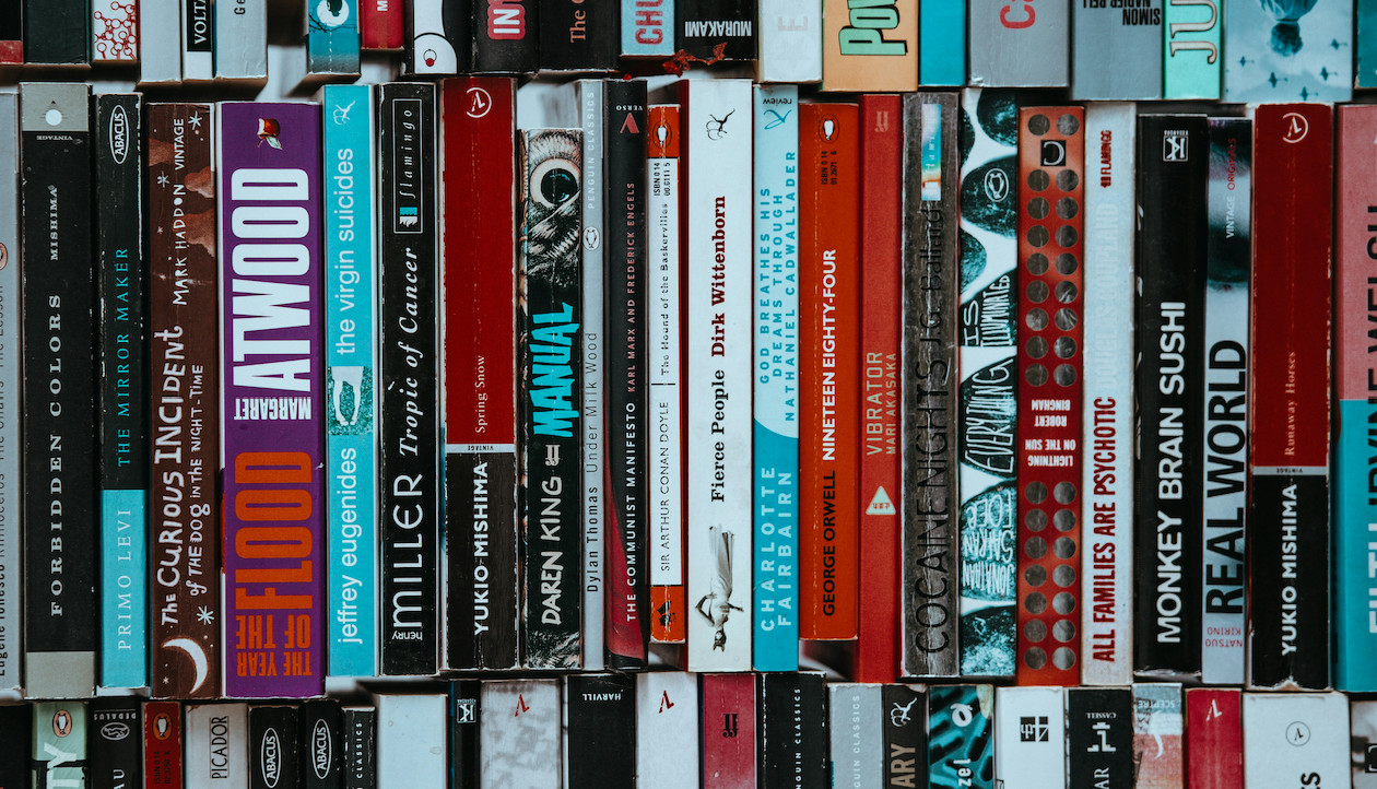 Un primo piano di numerosi libri impilati verticalmente e orizzontalmente su uno scaffale, che mostrano vari dorsi colorati con titoli, autori e disegni diversi.