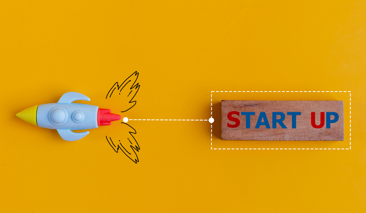 Un razzo giocattolo con le ali disegnate punta verso un blocco di legno con la scritta "START UP" su uno sfondo giallo brillante, a simboleggiare il lancio di una nuova attività finanziata dal capitale di rischio.