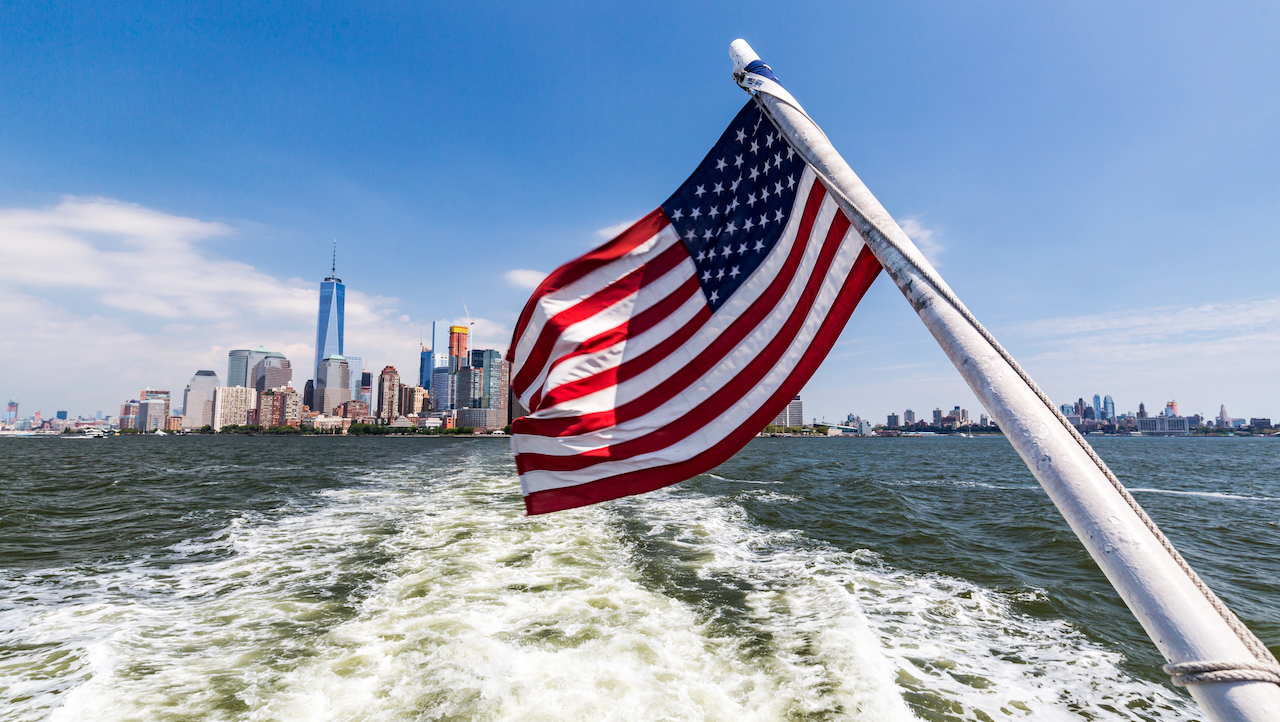 La bandiera americana sventola su una barca in acqua, con lo skyline di New York e il One World Trade Center visibili sullo sfondo sotto un cielo azzurro: un simbolo di resilienza in mezzo all'inflazione statunitense.
