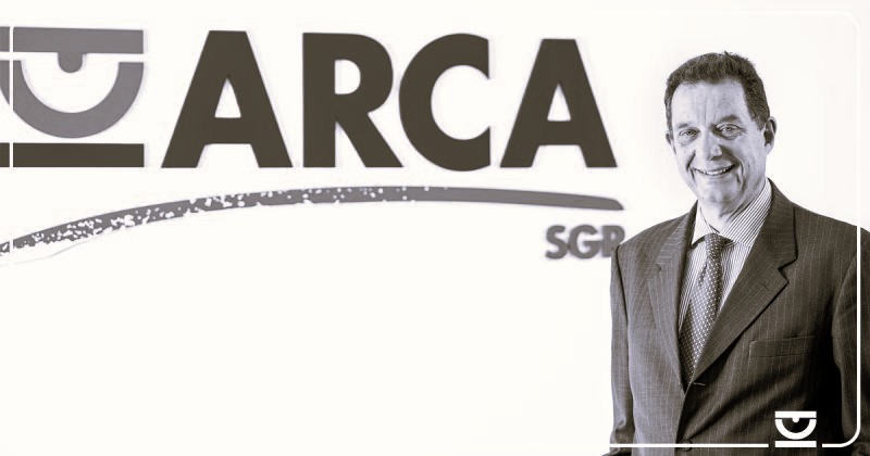 Una persona in giacca e cravatta è in piedi davanti a un muro con la scritta "ARCA SGR". L'immagine è in bianco e nero.
