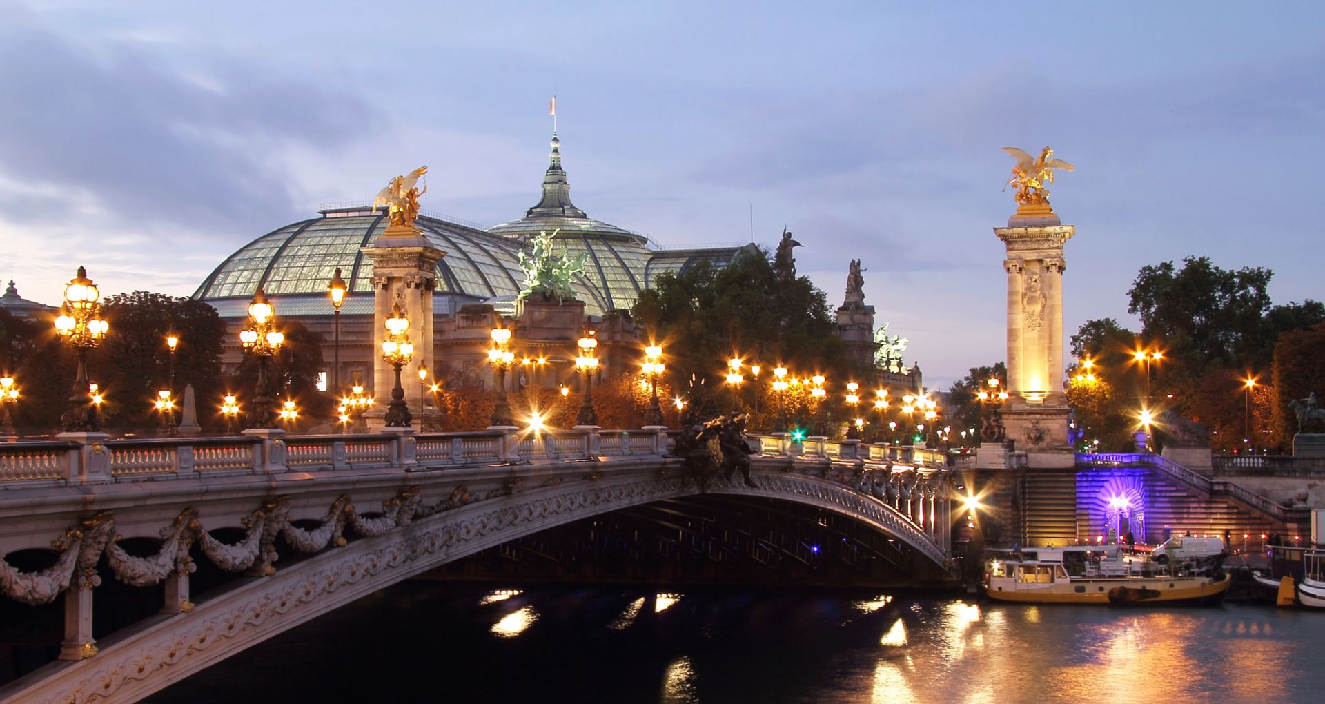 Una vista di Parigi al tramonto che mostra l'ornato ponte Pont Alexandre III adornato da lampioni e sculture decorative. Il Grand Palais è visibile sullo sfondo con il suo tetto a cupola di vetro. La Senna riflette le luci della città.