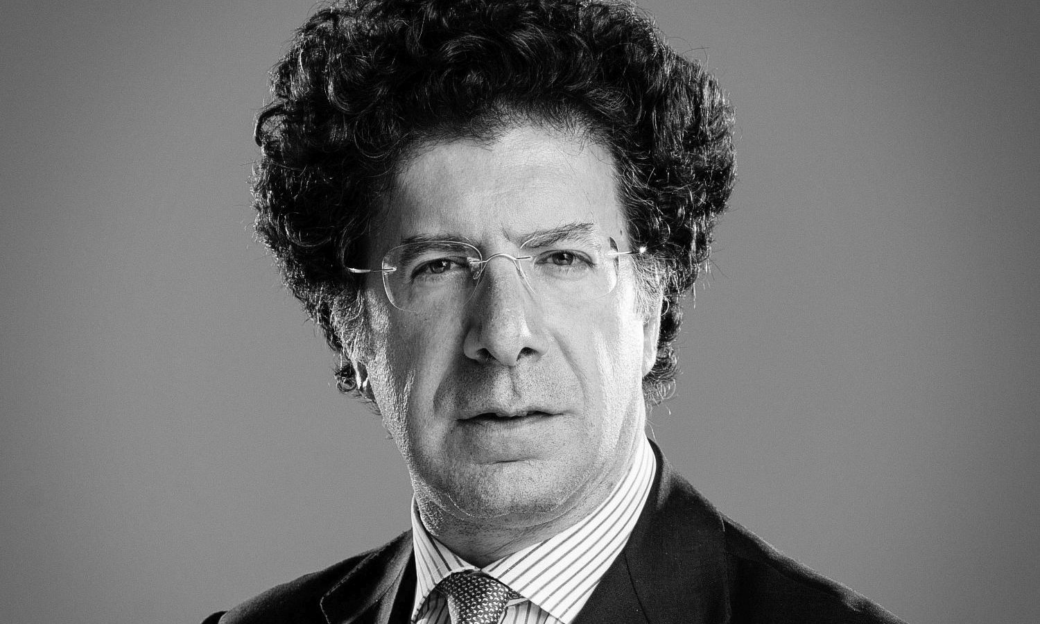 Ritratto in bianco e nero di un uomo con i capelli ricci e gli occhiali, che indossa un abito e una camicia a righe che accenna a un tocco di stile moderno ispirato a Monet e parla delle tasse pagate su questo quadro. Guarda dritto nella telecamera, su uno sfondo neutro che ricorda lo studio riflessivo di un artista.
