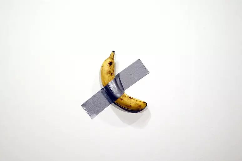 Una banana viene attaccata a una parete bianca con un unico pezzo di nastro adesivo grigio, posizionato in diagonale al centro del frutto.