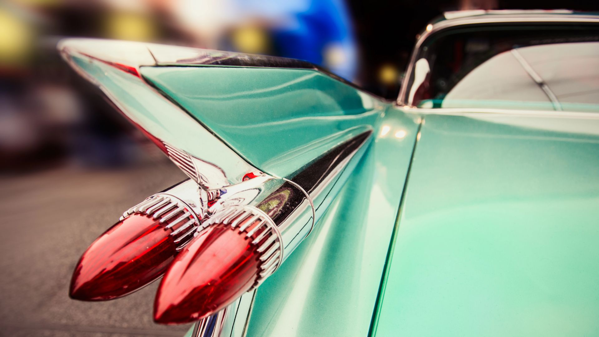 Primo piano della pinna posteriore verde acqua di un'auto d'epoca e dei fanali posteriori a forma di proiettile rosso, che ne mettono in mostra il design classico. Lo sfondo è sfocato, evidenziando i dettagli cromati lucidi e i colori audaci dell'auto.
