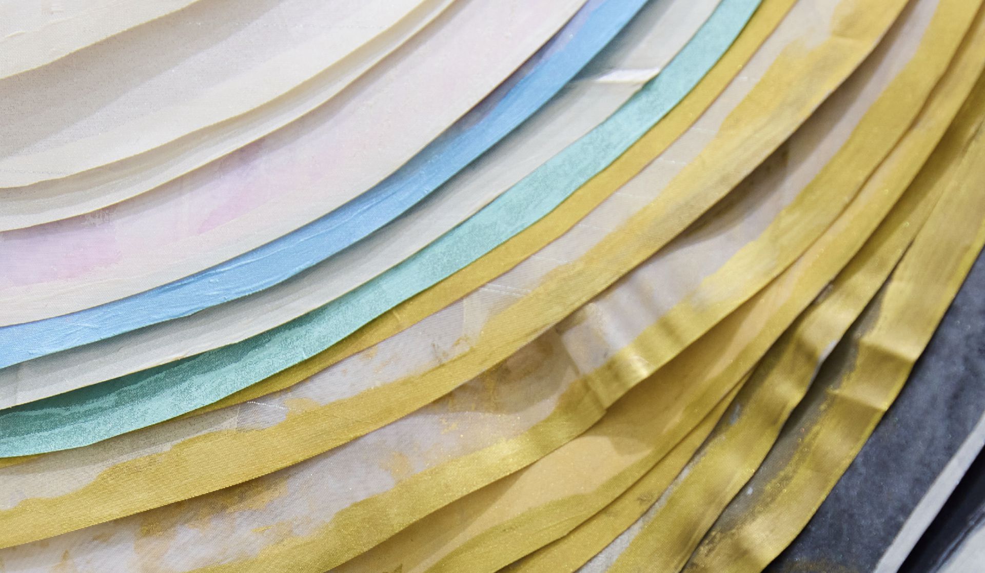 Primo piano di carta traslucida a strati in vari colori, tra cui blu, verde, giallo e grigio, disposti in un motivo curvo. La texture è leggermente ondulata, con la luce che si riflette sulla superficie.