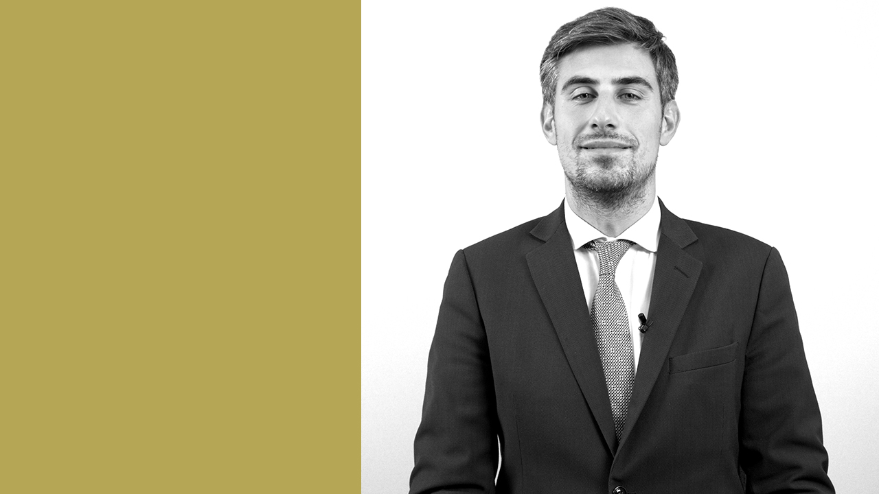 Un uomo in giacca e cravatta è in piedi su uno sfondo diviso, con un colore verde chiaro a sinistra e bianco a destra. Ha i capelli corti e la barba e guarda con sicurezza la telecamera. L'immagine è in bianco e nero.